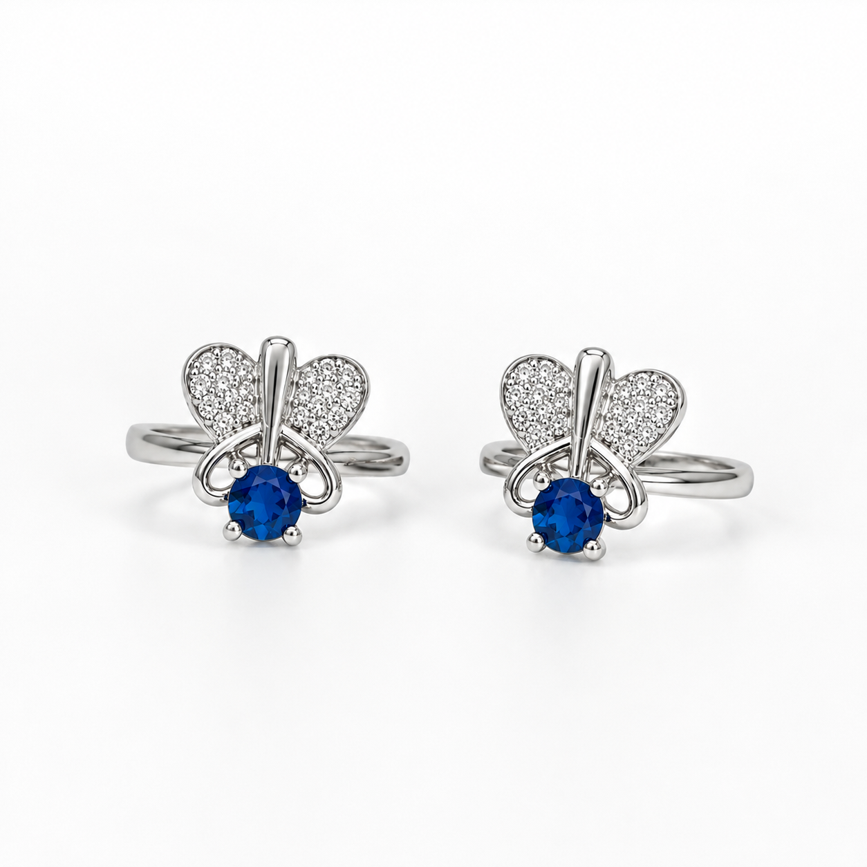 A pair of silver butterfly stud toe rings, each featuring a round blue sapphire at the center and pavé-set clear stones in the wings, shown on a clean white background.