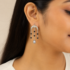 A close-up of a model's ear wearing a dangling earring. The earring features a silver crescent-shaped top with small clear stones, three small black beads, and a teardrop-shaped silver pearl at the bottom.