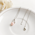 A delicate silver necklace with a single light pink pearl and a small clear crystal accent, displayed on a white ceramic plate with a white flower in the background.