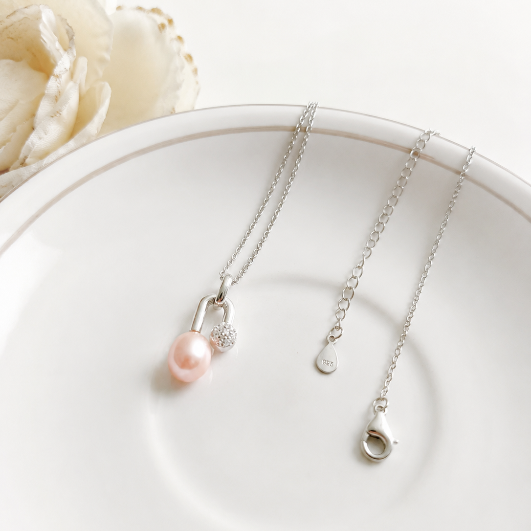 A delicate silver necklace with a single light pink pearl and a small clear crystal accent, displayed on a white ceramic plate with a white flower in the background.