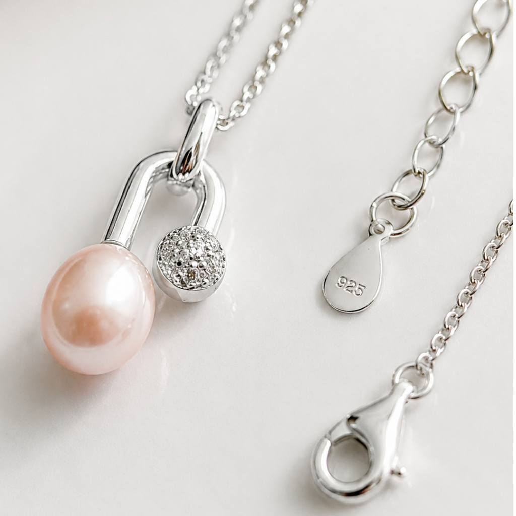 Silver pendant necklace featuring a light pink freshwater pearl and a small crystal-accented charm, displayed on a clean white surface with close-up views of the clasp and chain.
