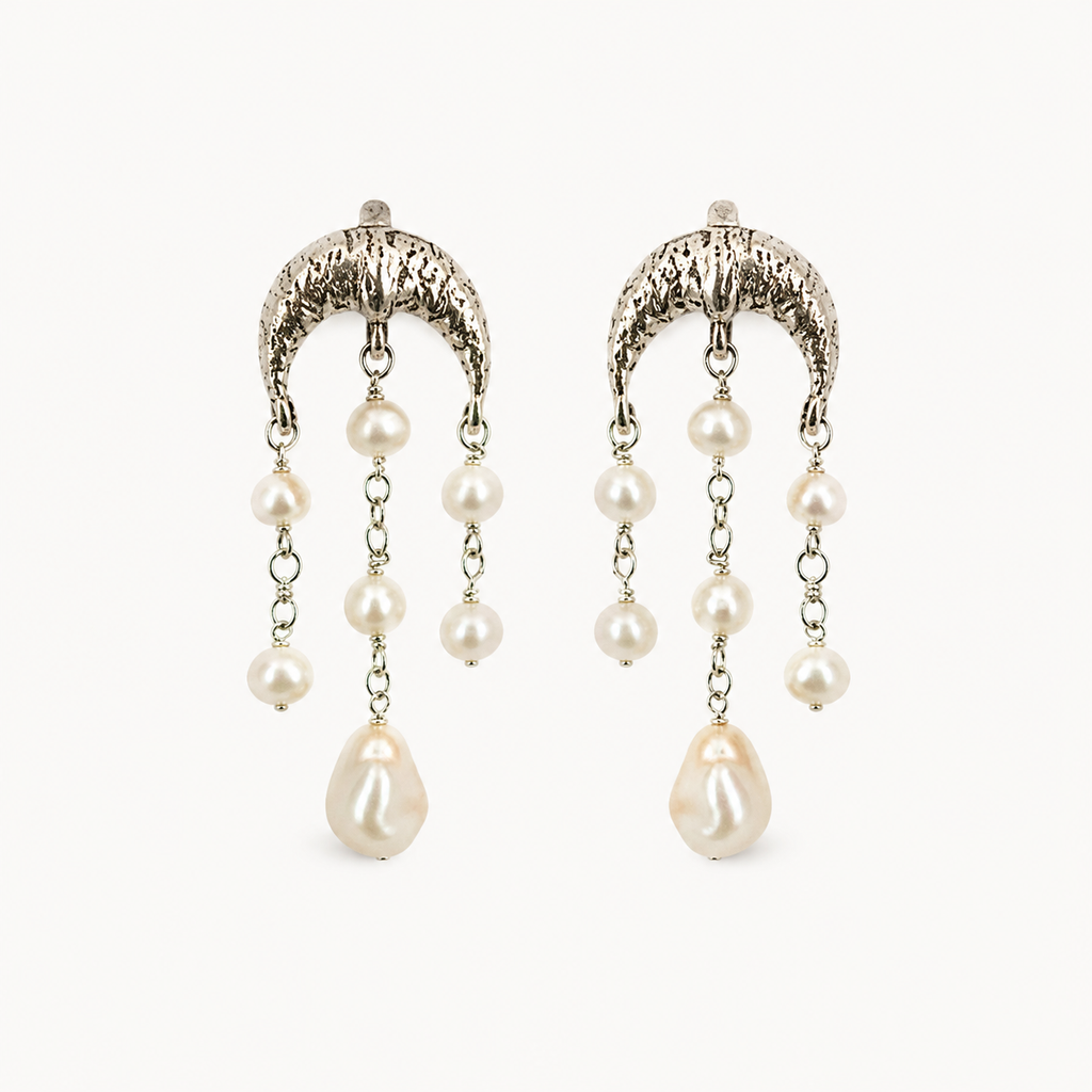 A pair of silver-toned dangle earrings with textured crescent-shaped tops, small pearl-like beads on chains, and larger teardrop pearl drops, shown on a plain light background.