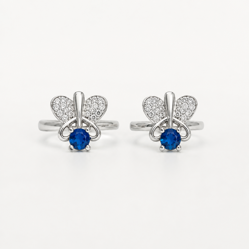 A pair of silver-tone toe rings featuring a butterfly design with pavé-set clear stones and a central round blue gemstone, shown on a plain light background.