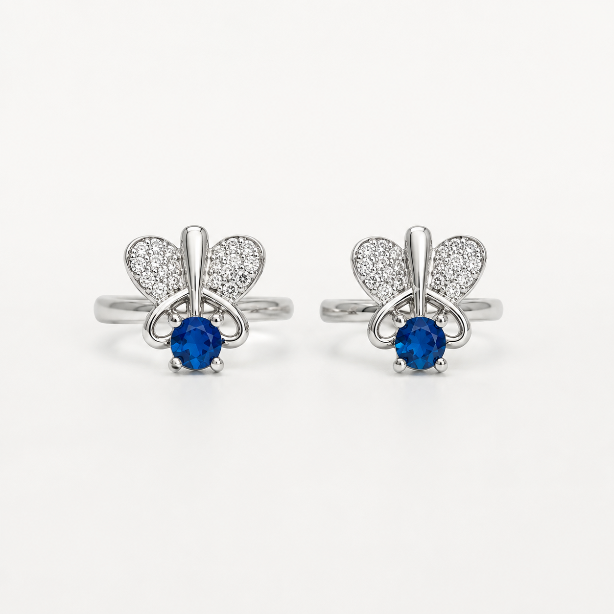 A pair of silver-tone toe rings featuring a butterfly design with pavé-set clear stones and a central round blue gemstone, shown on a plain light background.