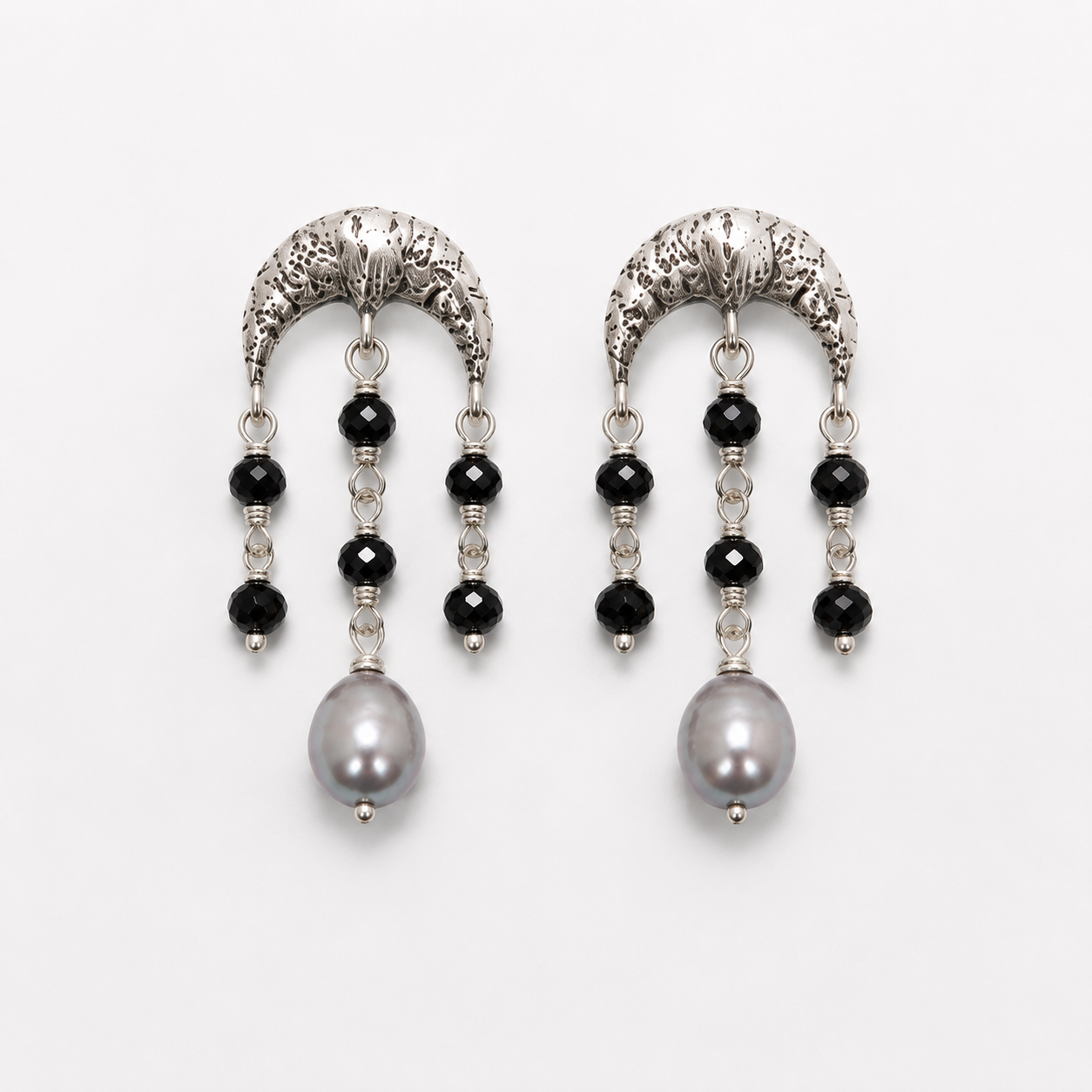 A pair of silver crescent-shaped dangle earrings with black faceted beads, clear spacer beads, and a large gray pearl at the bottom, shown on a light gray background.