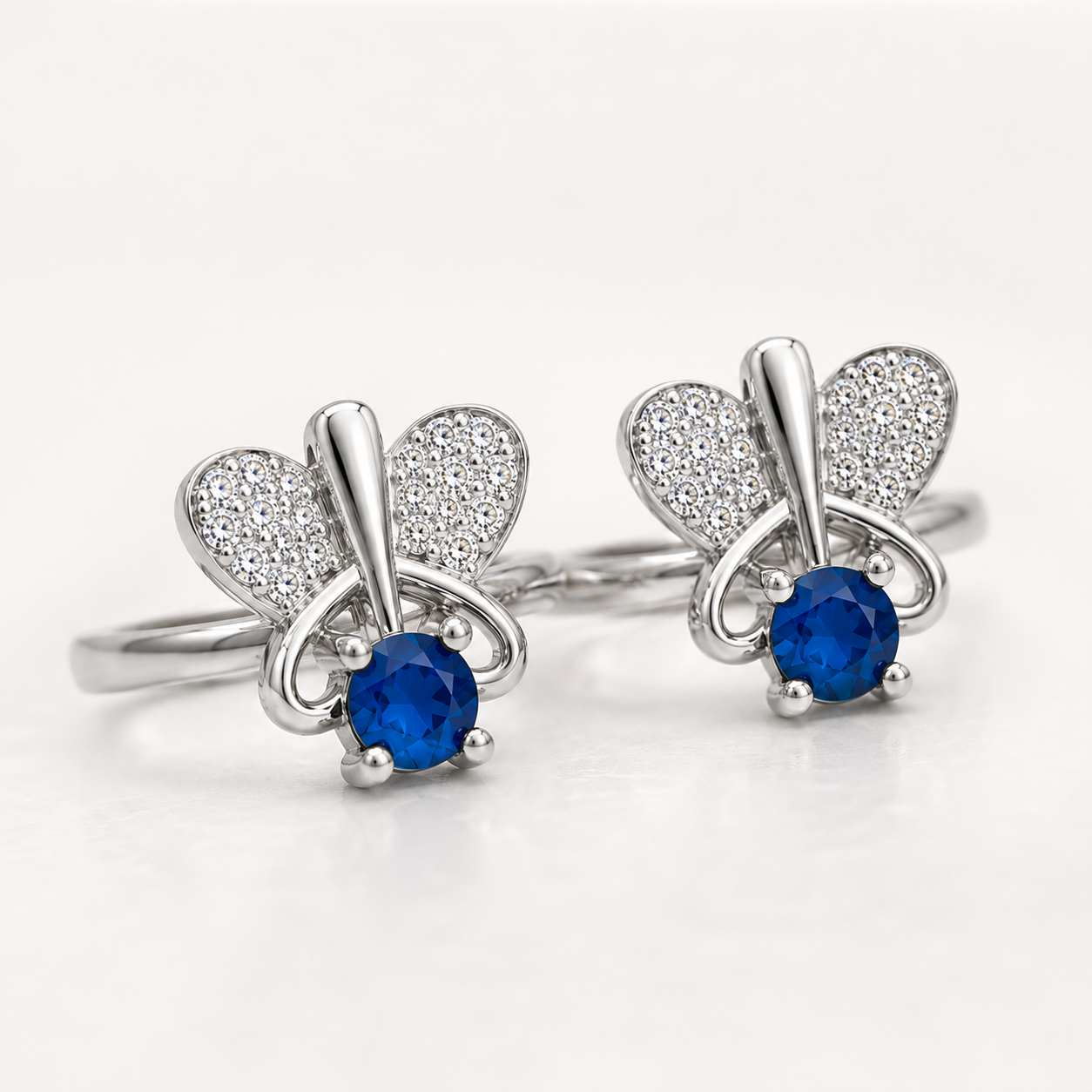 A pair of silver butterfly-shaped toe rings with pavé-set heart wings and a round blue gemstone at the center, shown on a clean white background.