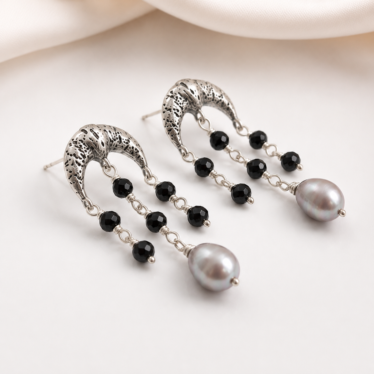 A pair of silver-toned dangle earrings with textured crescent-shaped tops, small black faceted beads, and a large gray pearl drop, photographed on a soft white background.