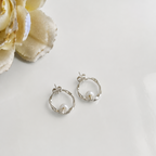 A pair of silver hoop earrings with a small white pearl inside each hoop, displayed on a clean white surface next to a decorative cream-colored flower.