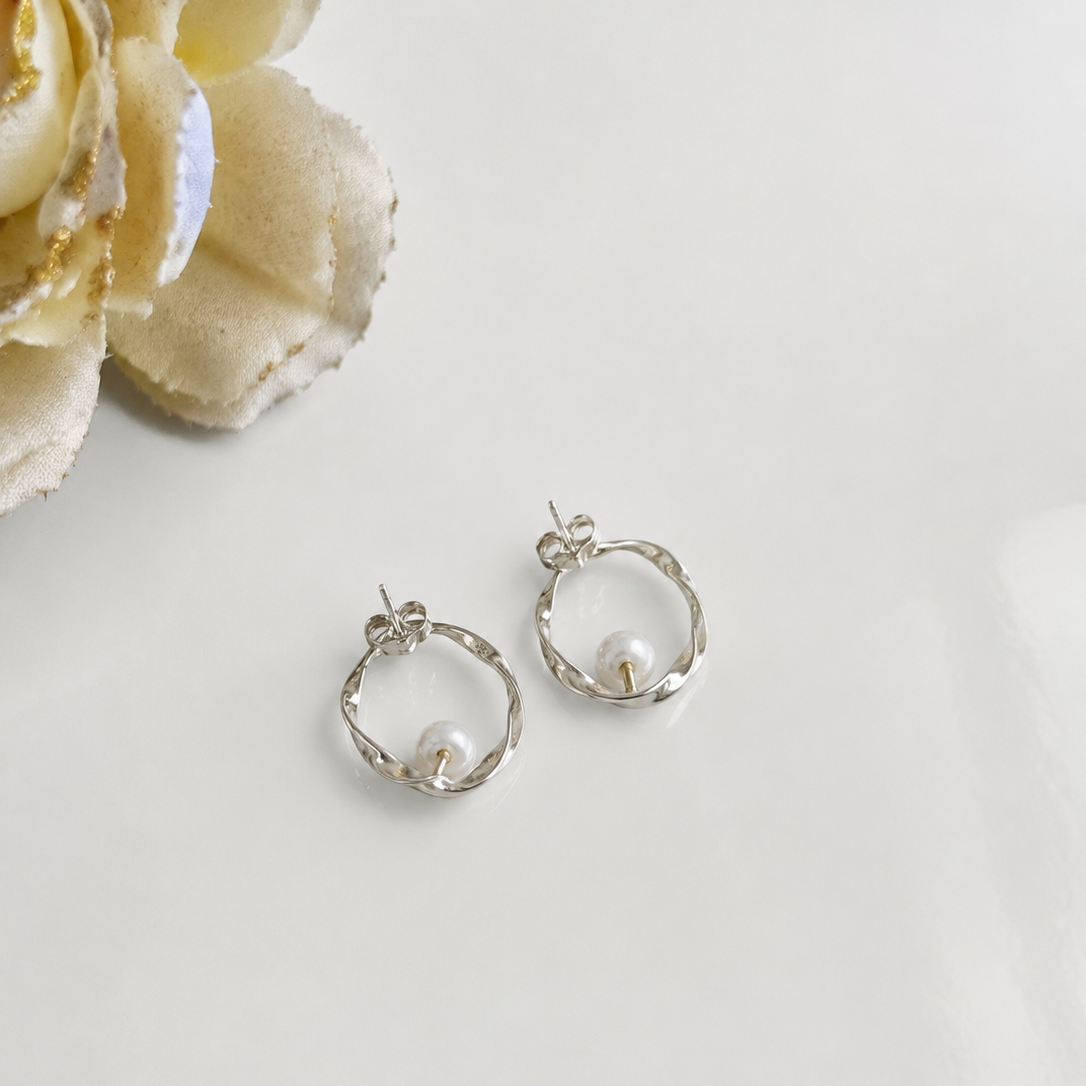 A pair of silver hoop earrings with a small white pearl inside each hoop, displayed on a clean white surface next to a decorative cream-colored flower.