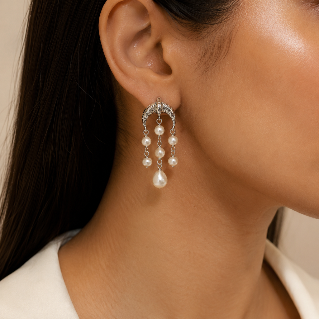 A close-up of a model's ear wearing a silver crescent-shaped dangle earring adorned with small clear  dangling white pearls.