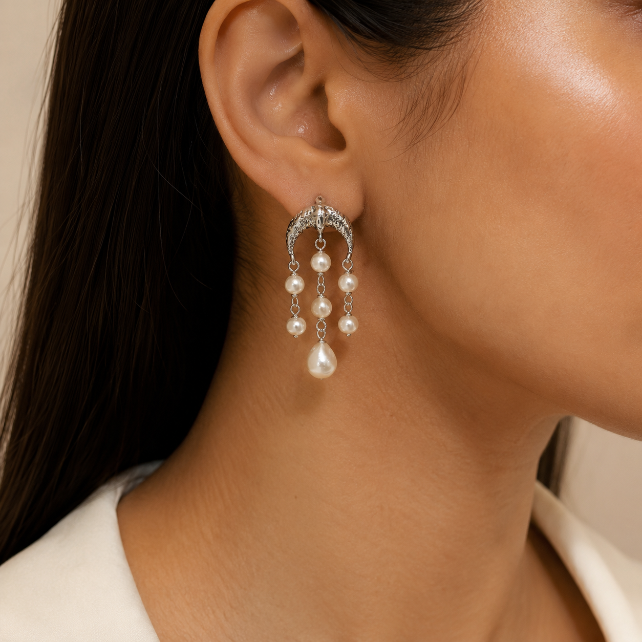 A close-up of a model's ear wearing a silver crescent-shaped dangle earring adorned with small clear  dangling white pearls.