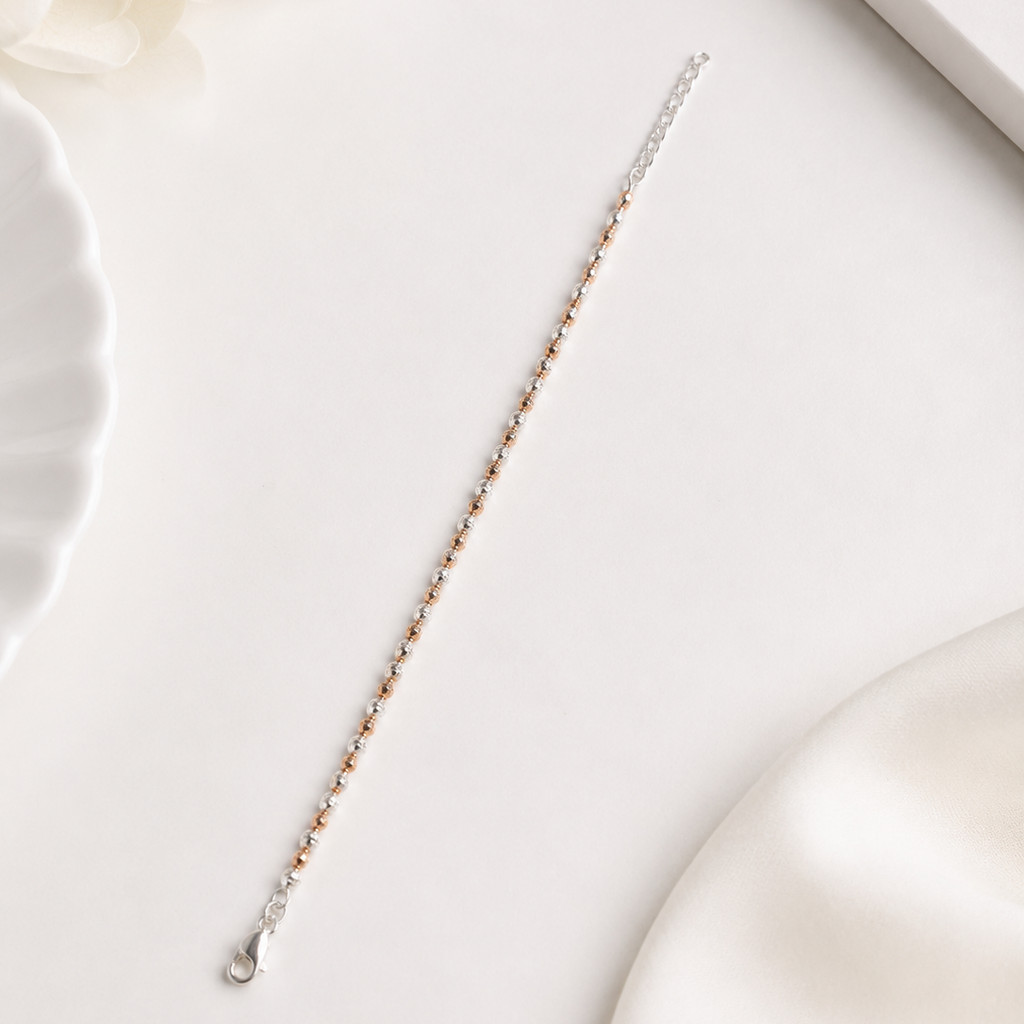 A delicate silver chain bracelet with small rose gold beads, laid diagonally on a clean white surface with soft decorative props.
