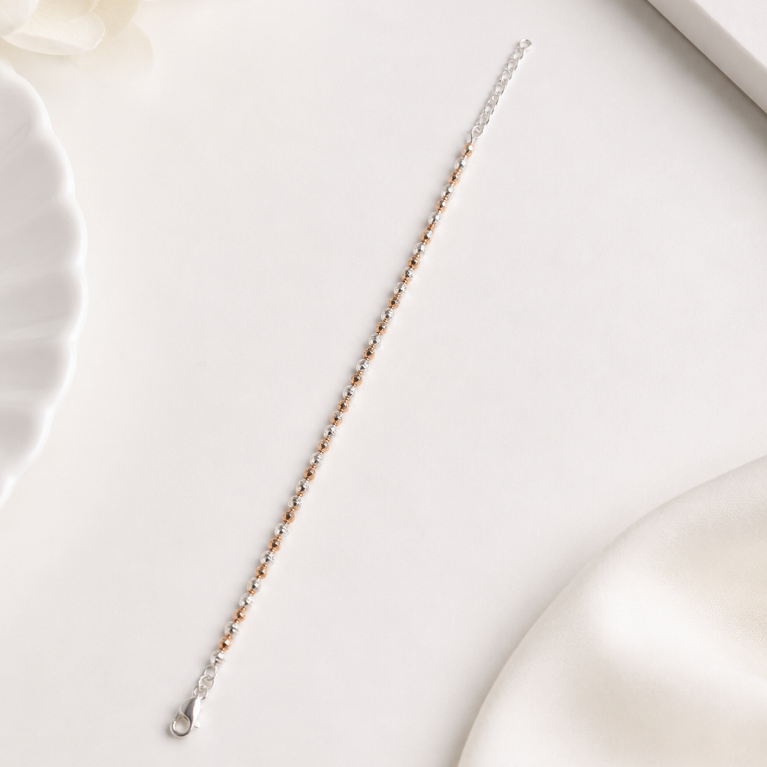 A delicate silver chain bracelet with small rose gold beads, laid diagonally on a clean white surface with soft decorative props.
