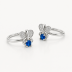 A pair of silver toe rings featuring a butterfly design with a blue oval gemstone and pavé-set heart wings, shown on a clean white background.