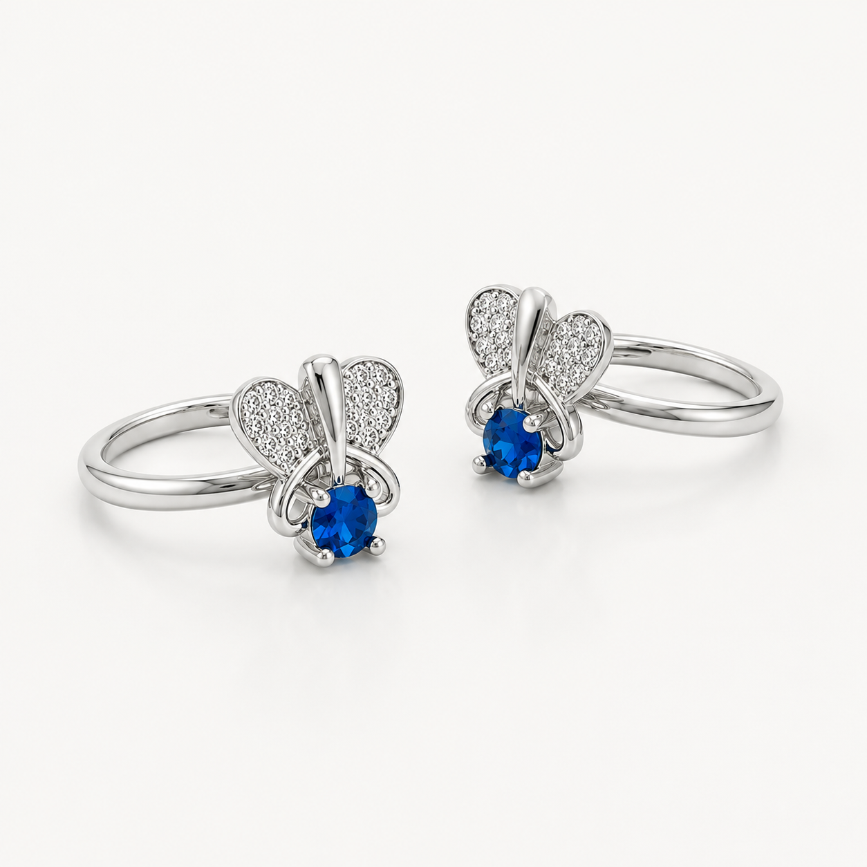 A pair of silver toe rings featuring a butterfly design with a blue oval gemstone and pavé-set heart wings, shown on a clean white background.