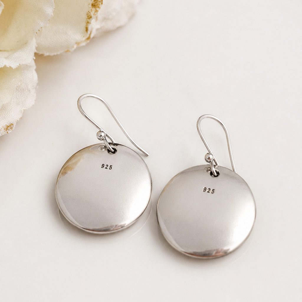 A pair of sterling silver disc drop earrings with fishhook wires, featuring a small '925' silver purity stamp on each disc. The earrings are displayed on a clean, light off-white surface next to a decorative white flower.