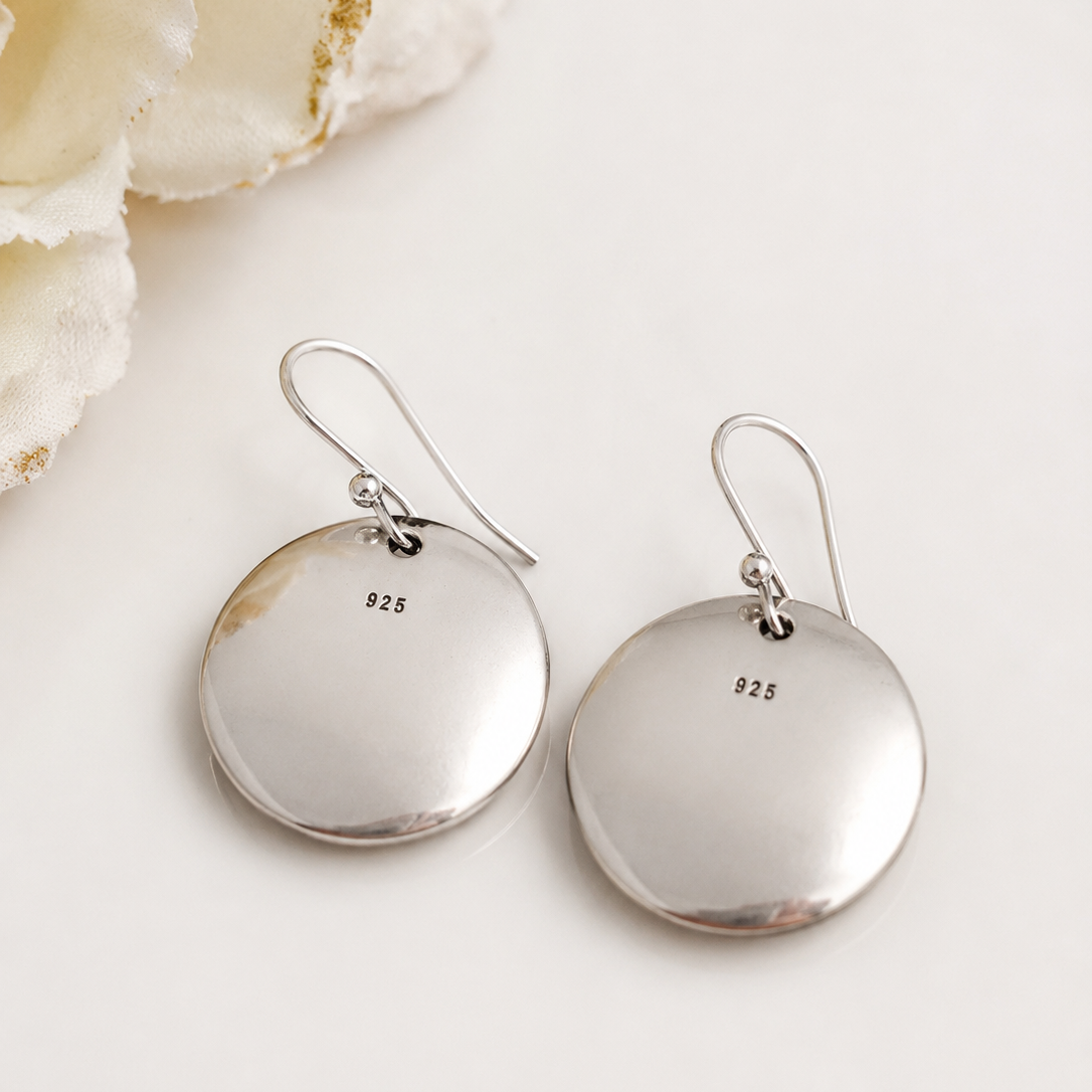 A pair of sterling silver disc drop earrings with fishhook wires, featuring a small '925' silver purity stamp on each disc. The earrings are displayed on a clean, light off-white surface next to a decorative white flower.
