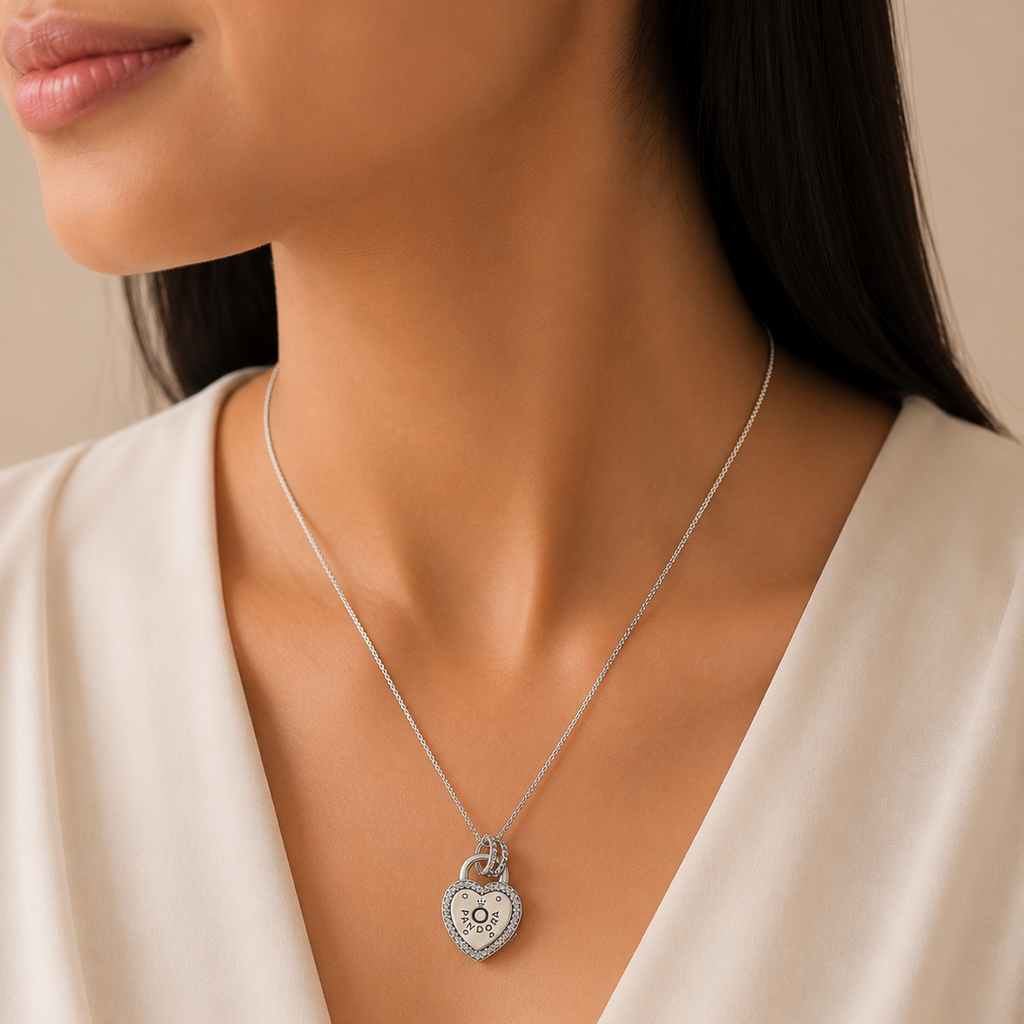 A close-up of a woman wearing a delicate silver heart-shaped pendant necklace with a central monogram design, paired with a cream-colored V-neck top.