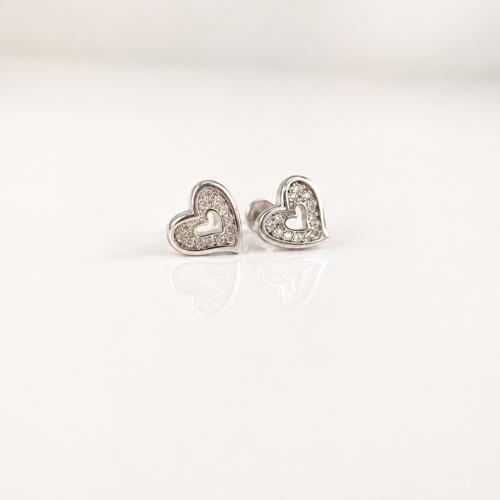 A pair of silver heart stud earrings with clear crystal accents, displayed on a clean white reflective surface.