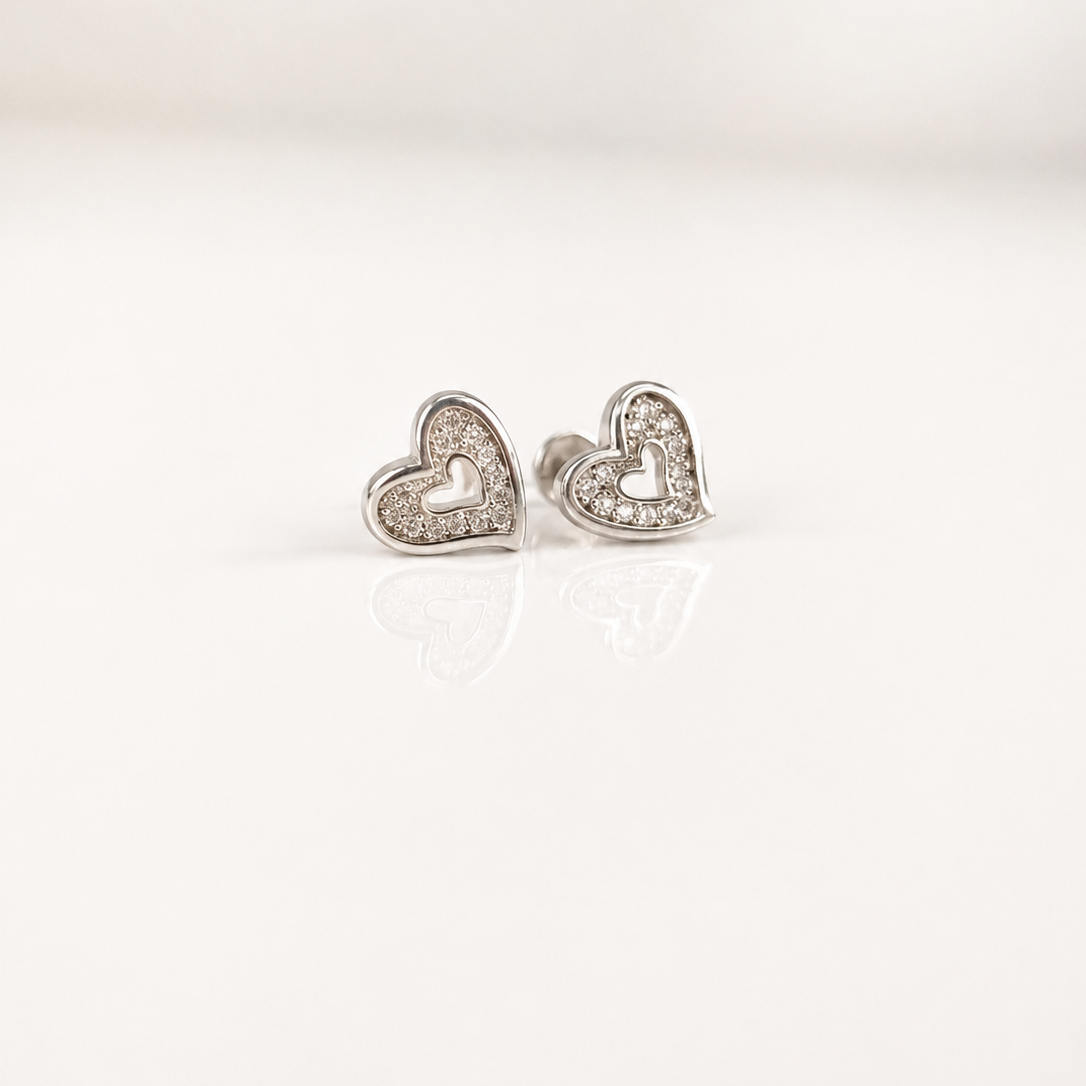 A pair of silver heart stud earrings with clear crystal accents, displayed on a clean white reflective surface.